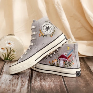 May include: Gray high-top Converse sneakers with embroidered floral and mushroom designs. The sneakers have a white rubber sole and a black Converse All Star logo.