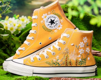 Può includere: Un paio di scarpe da ginnastica Converse alte gialle con fiori ricamati e un design a ragnatela. Le scarpe hanno una suola in gomma bianca e cuciture nere. La scarpa sinistra ha il logo Converse e il testo "Chuck Taylor All Star".