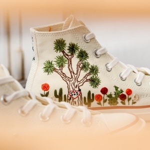 May include: White high-top canvas sneakers with embroidered Joshua Tree and red flowers. The sneakers have a white rubber sole and white laces. The embroidery includes the initials "S+S" in a heart.