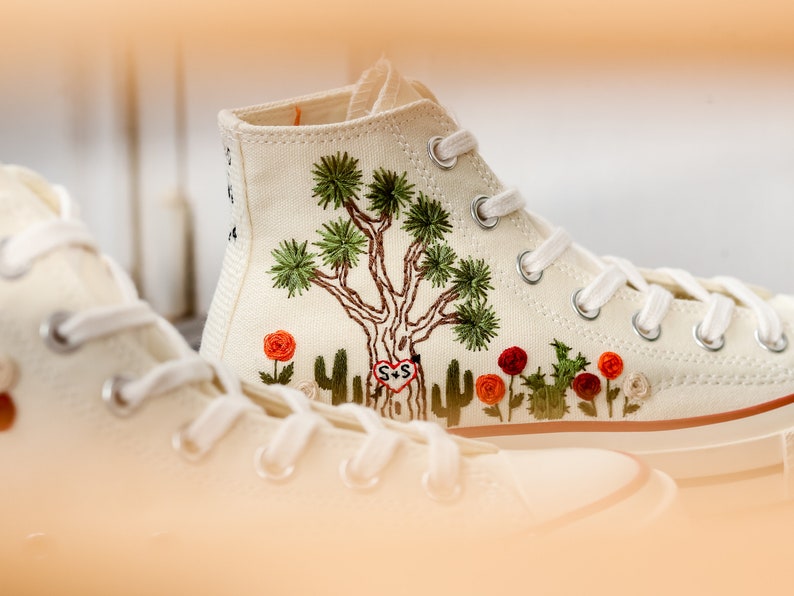 May include: White high-top canvas sneakers with embroidered Joshua Tree and red flowers. The sneakers have a white rubber sole and white laces. The embroidery includes the initials "S+S" in a heart.