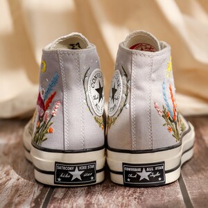 May include: A pair of gray canvas high-top sneakers with embroidered floral designs. The sneakers have black soles and white stitching. The left shoe has the text "SATRCONY HIKE STAR" on the sole. The right shoe has the text "CONVERSE ALL STAR Chuck Taylor" on the sole.