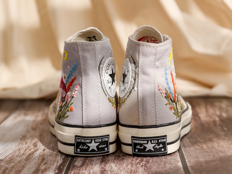 May include: A pair of gray canvas high-top sneakers with embroidered floral designs. The sneakers have black soles and white stitching. The left shoe has the text "SATRCONY HIKE STAR" on the sole. The right shoe has the text "CONVERSE ALL STAR Chuck Taylor" on the sole.