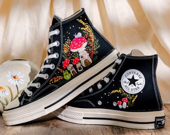 May include: Black high-top sneakers with white laces and soles. The sneakers feature embroidered mushroom and floral designs in red, yellow, green, and brown. The Converse logo is visible on the side.