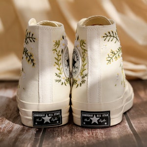 May include: A pair of white high-top canvas sneakers with floral embroidery. The sneakers have black laces and a black "Converse All Star" logo on the side.