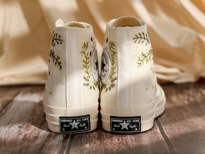 May include: A pair of white high-top canvas sneakers with floral embroidery. The sneakers have black laces and a black "Converse All Star" logo on the side.