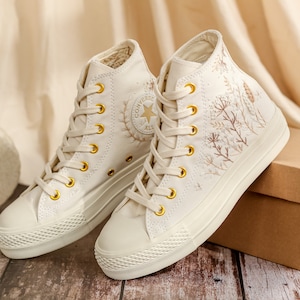 May include: A pair of white high-top trainers with embroidered floral designs and gold accents. The trainers are on a wooden surface with a brown box in the background.