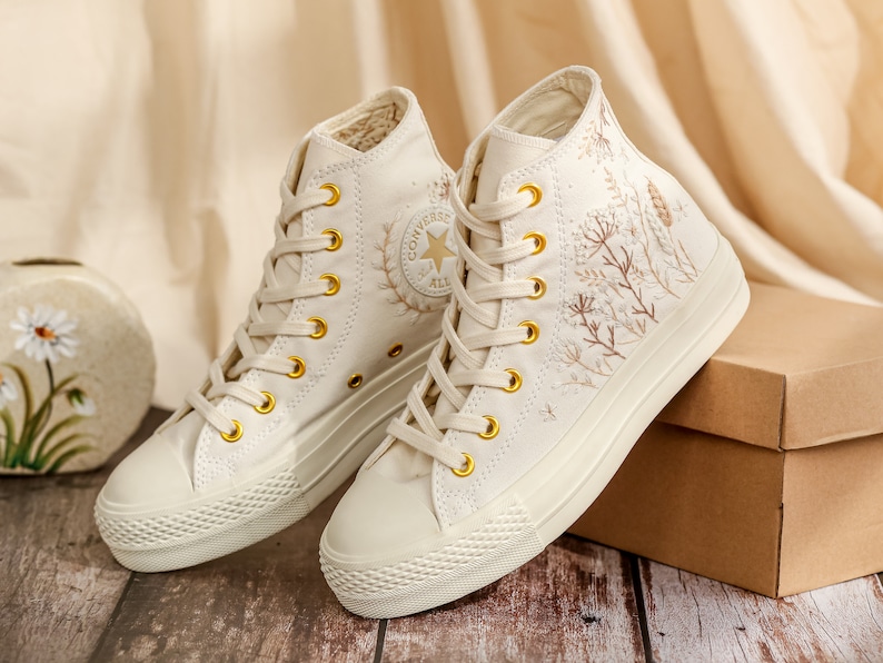 May include: A pair of white high-top trainers with embroidered floral designs and gold accents. The trainers are on a wooden surface with a brown box in the background.
