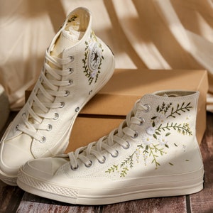 May include: A pair of off-white high-top canvas sneakers with embroidered floral designs. The sneakers have white laces and a white rubber sole.