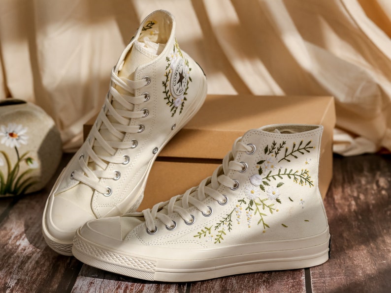 May include: A pair of off-white high-top canvas sneakers with embroidered floral designs. The sneakers have white laces and a white rubber sole.