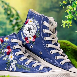 May include: A pair of blue high-top Converse trainers with white laces and embroidered floral designs. The Converse All Star logo is visible on the side of the shoe.