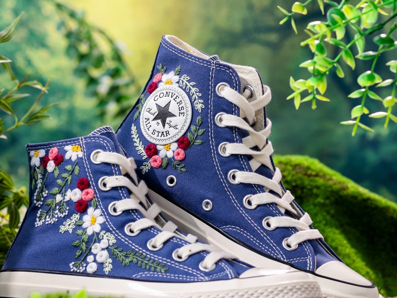 May include: A pair of blue high-top Converse trainers with white laces and embroidered floral designs. The Converse All Star logo is visible on the side of the shoe.