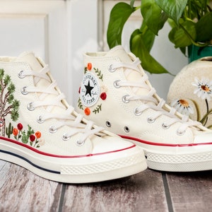 May include: A pair of white high-top canvas sneakers with red trim and embroidered floral designs. The sneakers have white laces and a black star logo on the side.