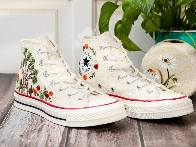 May include: A pair of white high-top canvas sneakers with red trim and embroidered floral designs. The sneakers have white laces and a black star logo on the side.