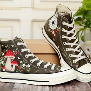 May include: A pair of olive green high-top canvas trainers with white laces and a white rubber sole. The trainers are embroidered with red and white mushrooms, green leaves, and white flowers.