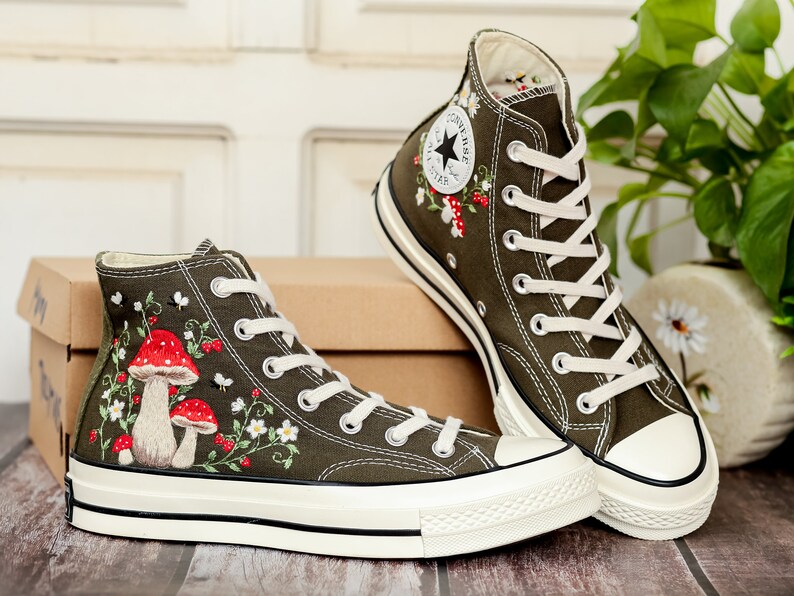 May include: A pair of olive green high-top canvas trainers with white laces and a white rubber sole. The trainers are embroidered with red and white mushrooms, green leaves, and white flowers.