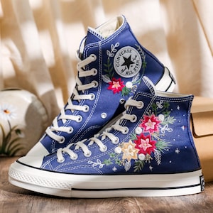 Puede incluir: Un par de zapatillas de lona azul de caña alta con cordones blancos y suela de goma blanca. Las zapatillas tienen flores bordadas en rojo, rosa y amarillo en los laterales. Las zapatillas tienen el logotipo de Converse All Star en el lateral.