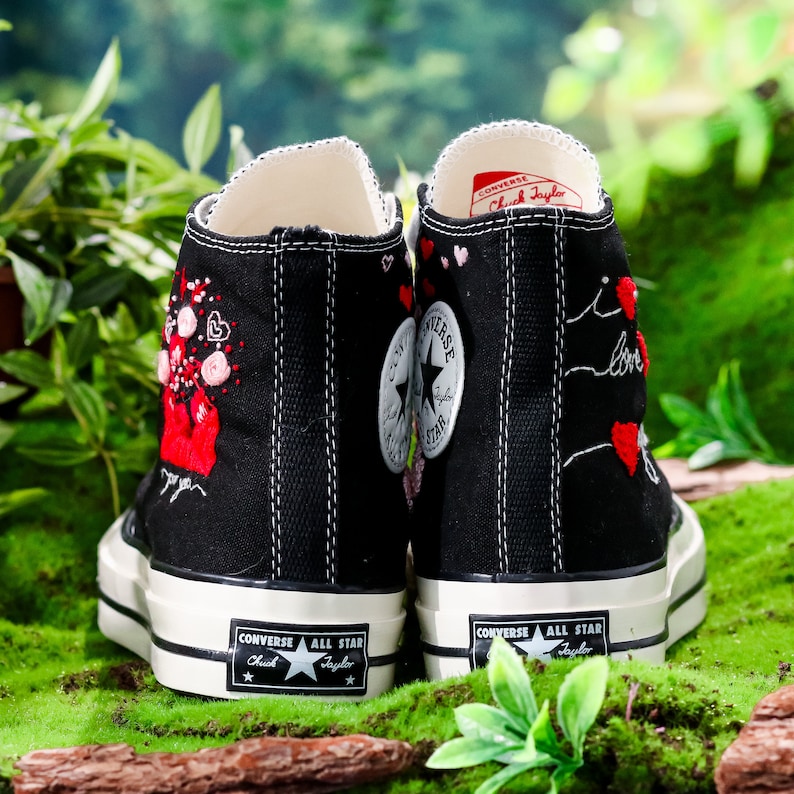 Op de afbeelding: Zwarte Converse sneakers met hoge schacht, witte zolen en rode geborduurde harten en bloemen. De tekst "Converse All Star Chuck Taylor" staat aan de zijkant van elke schoen.