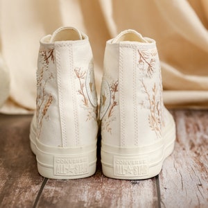 May include: A pair of off-white high-top canvas sneakers with brown floral embroidery and the text "Converse All Star" on the back.