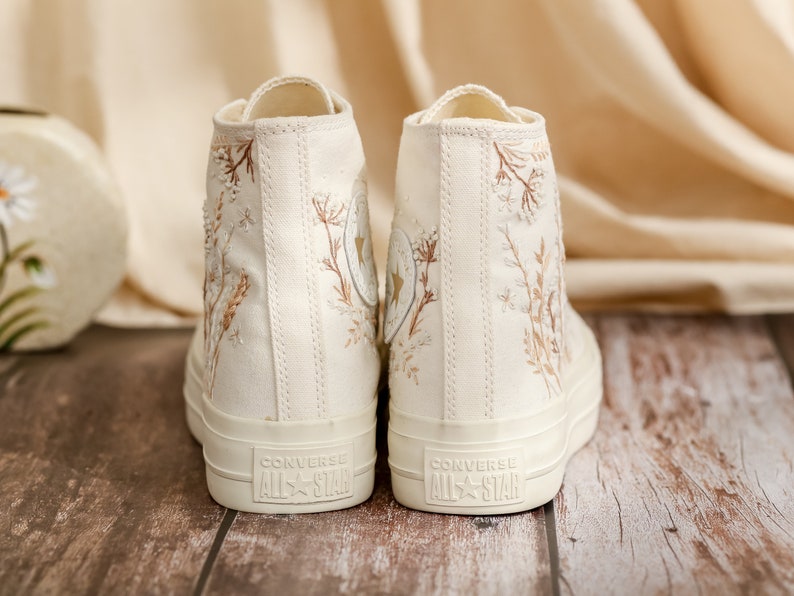 May include: A pair of off-white high-top canvas sneakers with brown floral embroidery and the text "Converse All Star" on the back.