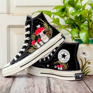 May include: A pair of black high-top Converse sneakers with white soles. The sneakers are decorated with embroidered mushrooms, leaves, and a snail. The left shoe has the Converse All Star logo on the side.