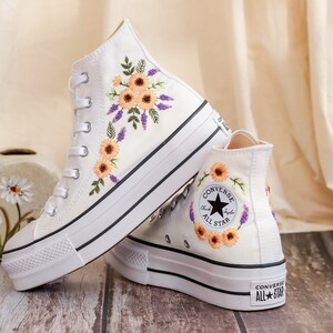 May include: Pair of white high-top sneakers with floral embroidery. The sneakers feature a platform sole, black trim, and a classic lace-up design. The embroidered flowers are in shades of orange, purple, and green.