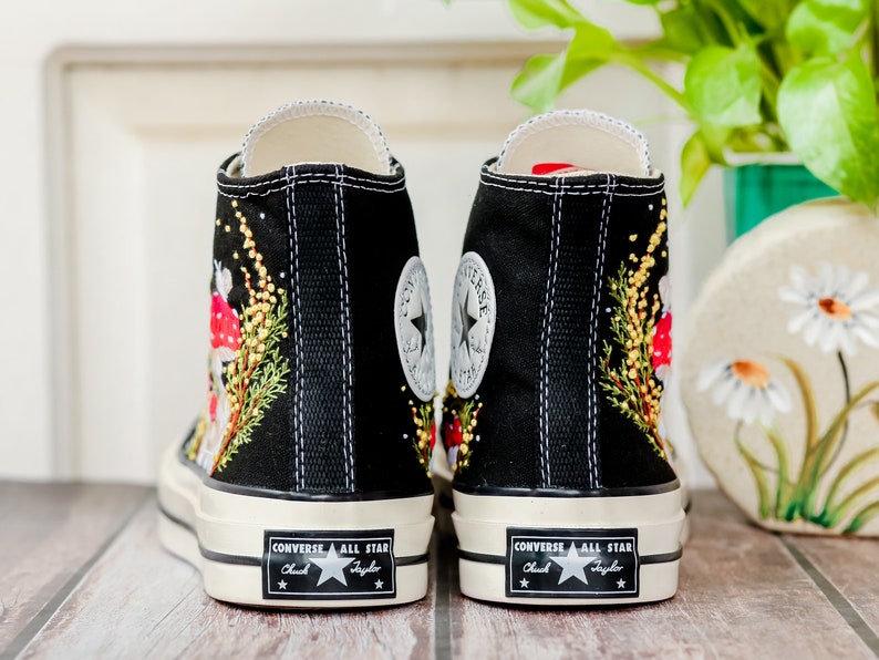 May include: Black high-top Converse sneakers with embroidered mushrooms and greenery. The sneakers have a white rubber sole and the Converse All Star logo is visible on the back.