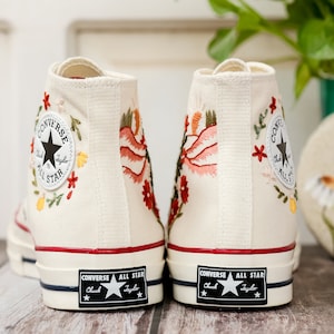 May include: A pair of white high-top Converse trainers with floral embroidery. The trainers have a red stripe around the top and a black Converse All Star logo on the back.
