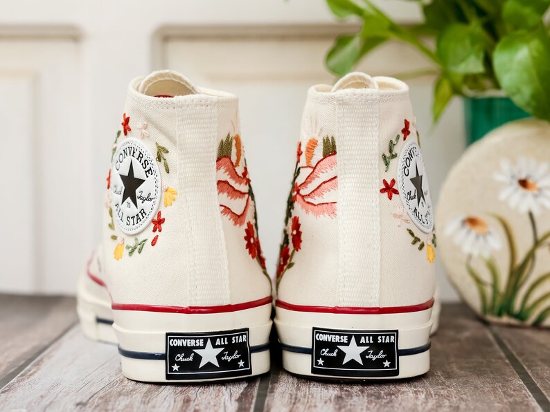 May include: A pair of white high-top Converse trainers with floral embroidery. The trainers have a red stripe around the top and a black Converse All Star logo on the back.