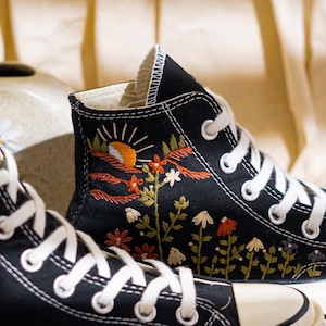 K&ouml;nnte beinhalten: Schwarze High-Top-Sneaker mit wei&szlig;en Schn&uuml;rsenkeln und einer cremefarbenen Zehenkappe. Die Sneaker haben farbenfrohe, gestickte Blumen- und Landschaftsdesigns, darunter eine Sonne, Blumen und Bl&auml;tter. Das Design befindet sich auf der Au&szlig;enseite des Schuhs.