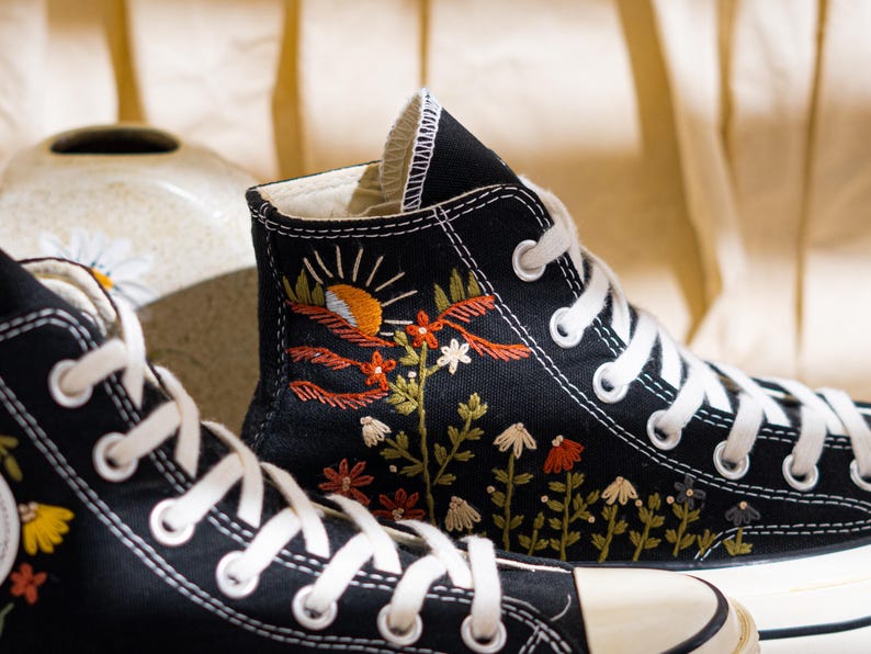 K&ouml;nnte beinhalten: Schwarze High-Top-Sneaker mit wei&szlig;en Schn&uuml;rsenkeln und einer cremefarbenen Zehenkappe. Die Sneaker haben farbenfrohe, gestickte Blumen- und Landschaftsdesigns, darunter eine Sonne, Blumen und Bl&auml;tter. Das Design befindet sich auf der Au&szlig;enseite des Schuhs.
