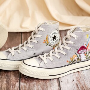 May include: Gray high-top Converse sneakers with white soles and embroidered floral and mushroom designs. The sneakers feature the Converse All Star logo on the side.