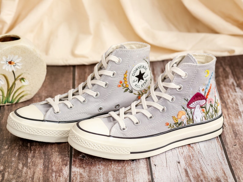 May include: Gray high-top Converse sneakers with white soles and embroidered floral and mushroom designs. The sneakers feature the Converse All Star logo on the side.