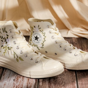 May include: A pair of white high-top Converse sneakers with floral embroidery and the Converse All Star logo. The sneakers are on a wooden surface.
