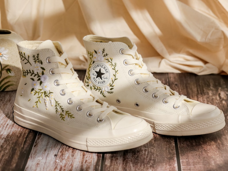 May include: A pair of white high-top Converse sneakers with floral embroidery and the Converse All Star logo. The sneakers are on a wooden surface.