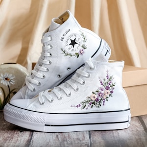 May include: White high-top Converse sneakers with a floral embroidery design on one shoe and the date "08.10.2024" and the Converse logo on the other shoe.
