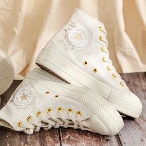 May include: A pair of white high-top Converse trainers with gold eyelets and a floral embroidery design. The Converse logo and star are visible on the side of the shoe.