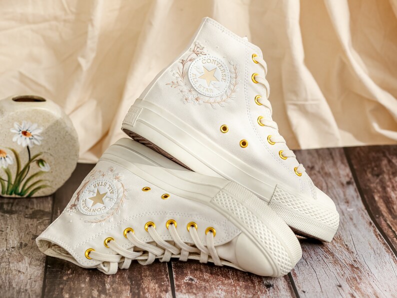May include: A pair of white high-top Converse trainers with gold eyelets and a floral embroidery design. The Converse logo and star are visible on the side of the shoe.