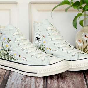 May include: A pair of light blue high-top canvas sneakers with white laces and a white rubber sole. The sneakers are embroidered with white flowers and green leaves.