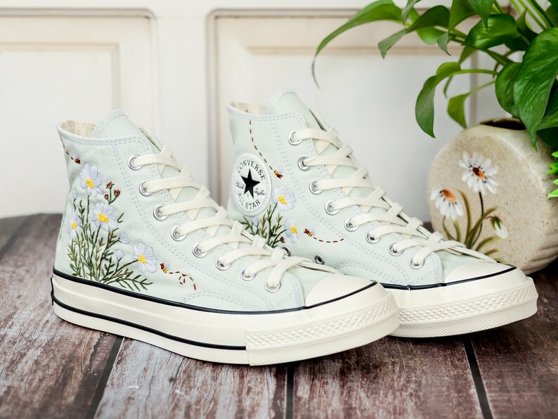 May include: A pair of light blue high-top canvas sneakers with white laces and a white rubber sole. The sneakers are embroidered with white flowers and green leaves.