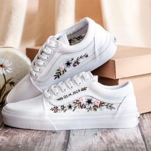 May include: White sneakers with floral embroidery in pink, green, and black. The shoes have white laces and soles. The date "09.14.2024" is embroidered on the side. The shoes are displayed on a wooden surface.