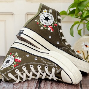 May include: A pair of olive green high-top Converse trainers with white soles and embroidered flowers and mushrooms. The trainers feature the Converse All Star logo on the side.