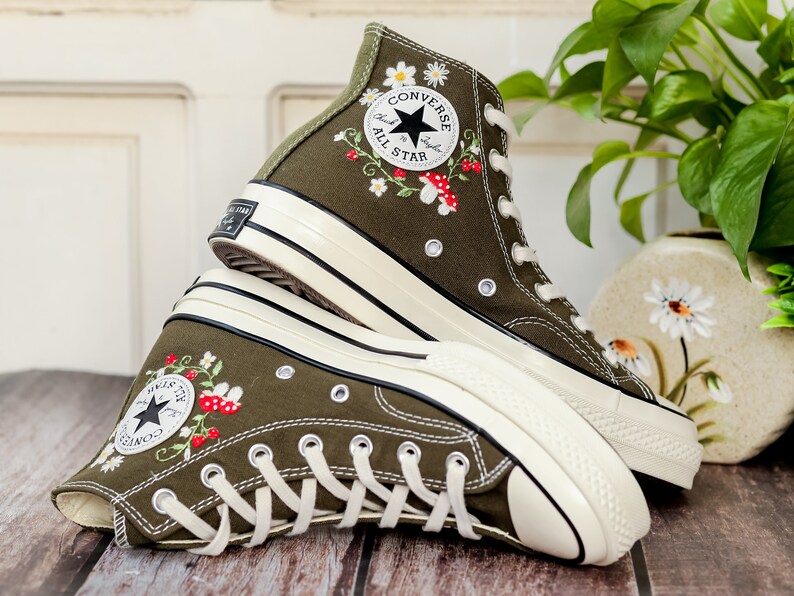 May include: A pair of olive green high-top Converse trainers with white soles and embroidered flowers and mushrooms. The trainers feature the Converse All Star logo on the side.
