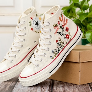 May include: A pair of white high-top canvas trainers with red trim and floral embroidery. The trainers feature a black star logo and the text "Converse All Star".