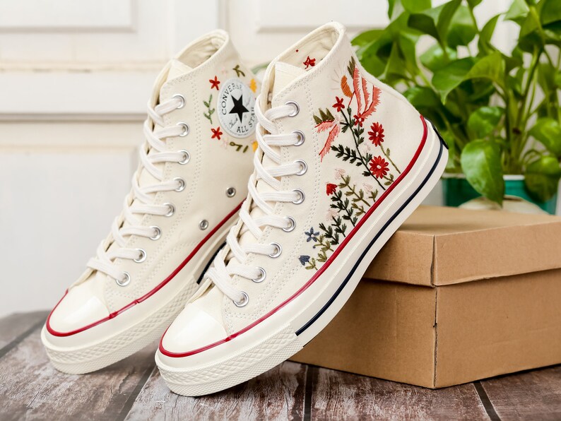 May include: A pair of white high-top canvas trainers with red trim and floral embroidery. The trainers feature a black star logo and the text "Converse All Star".
