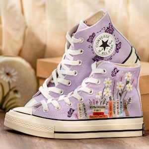 Può includere: Un paio di scarpe da ginnastica alte Converse Chuck Taylor All Star color lavanda. Le scarpe da ginnastica presentano lacci bianchi, una suola color crema e ricami floreali e a farfalla. Il logo Converse è visibile.