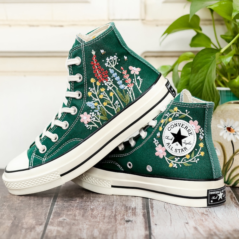 Może przedstawiać: Para zielonych, wysokich trampek z pł&oacute;ciennymi cholewkami i białymi podeszwami. Trampki są wyszywane kolorowymi kwiatami i mają czarne logo Converse All Star na boku.
