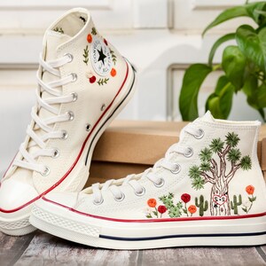 May include: A pair of white high-top canvas sneakers with red and green embroidered details. The sneakers have a white rubber sole and a white laces. One sneaker has a tree and cactus design, while the other has a floral design.