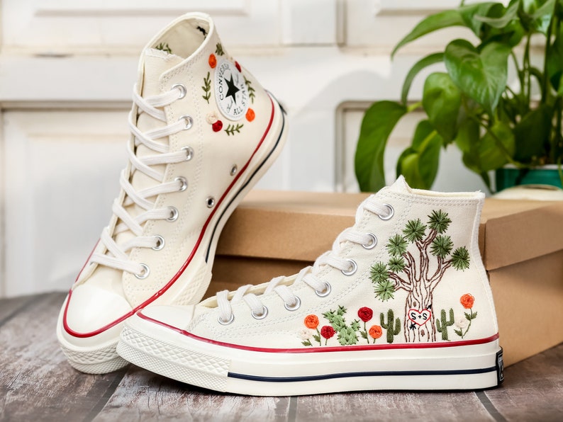 May include: A pair of white high-top canvas sneakers with red and green embroidered details. The sneakers have a white rubber sole and a white laces. One sneaker has a tree and cactus design, while the other has a floral design.