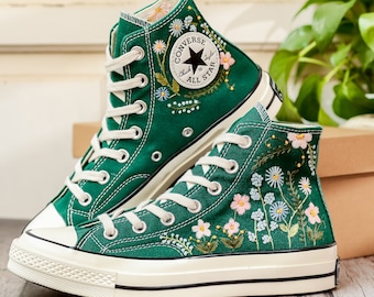 Può includere: Sneakers alte verdi con lacci bianchi e puntale in gomma bianca. Le sneakers presentano ricami floreali in rosa, bianco e blu. Il logo Converse All Star è visibile.
