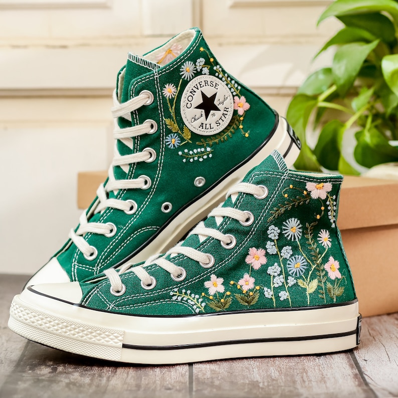 Puede incluir: Zapatillas altas verdes con cordones blancos y puntera de goma blanca. Las zapatillas presentan dise&ntilde;os florales bordados en rosa, blanco y azul. El logotipo de Converse All Star es visible.